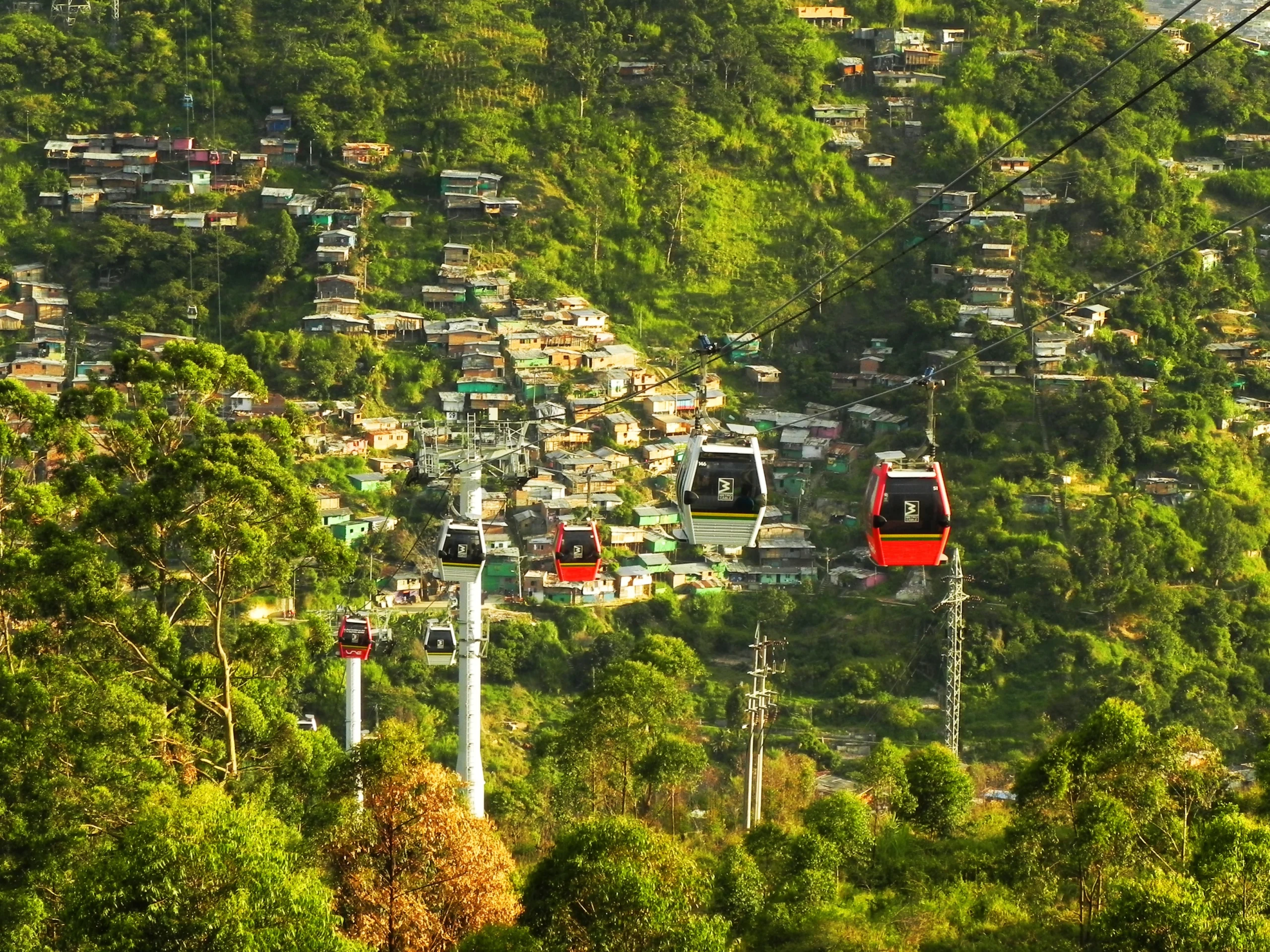 Medellín Metrocable gondolas above a hillside neighborhood in Colombia, with homes, trees, and steep green slopes.